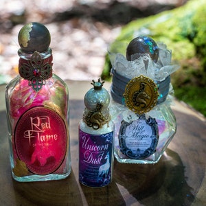 Puede incluir: Tres botellas de vidrio con etiquetas y tapones decorativos. Las botellas contienen purpurina de colores y est&aacute;n etiquetadas como "Red Flame of the Bull", "Unicorn Dust" y "The Will of Magic".