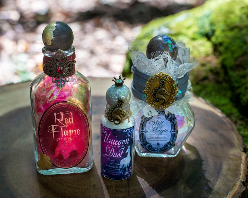 Puede incluir: Tres botellas de vidrio con etiquetas y tapones decorativos. Las botellas contienen purpurina de colores y est&aacute;n etiquetadas como "Red Flame of the Bull", "Unicorn Dust" y "The Will of Magic".