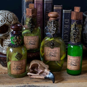 May include: Four decorative glass bottles filled with green liquid, labeled "Snake Oil." The bottles have ornate metal and fabric accents, wooden stoppers, and are arranged with a skull and books in the background. The bottles vary in size and shape.
