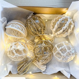 May include: A collection of six decorative glass ball ornaments with macrame detailing, presented in a cardboard box. The ornaments feature clear glass spheres encased in beige and off-white woven patterns. The box contains the text "OPTION 3".