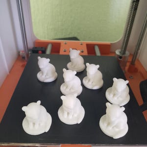 May include: Several small, white bear figurines, likely 3D-printed, are displayed on a black surface. The bears are depicted in a seated posture. The image is taken inside a 3D printer, with the word "CAUTION" visible.