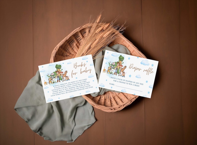 May include: Two baby shower invitation cards with dinosaur and animal illustrations. One card reads "Books for baby" and the other says "Diaper raffle". The cards are on a woven basket with wheat stalks and a sage green cloth.