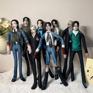 May include: A collection of seven fabric dolls featuring printed human faces and various outfits, including suits, jackets, and ties. The dolls have slender limbs and are arranged in a group, showcasing diverse clothing styles like denim, plaid, and solid colors.