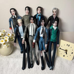 May include: A collection of eight fabric dolls featuring printed portraits of individuals in various outfits, including blazers, sweaters, and shirts. The dolls are arranged in a group, each with distinct facial features and clothing styles.