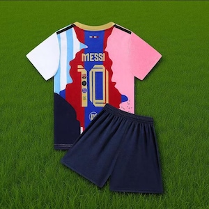Może przedstawiać: Dwuczęściowy zestaw stroju piłkarskiego na zielonym tle. Koszulka jest biała, niebieska, czerwona i różowa z napisem "MESSI" i numerem "10" z przodu. Spodenki są granatowe.