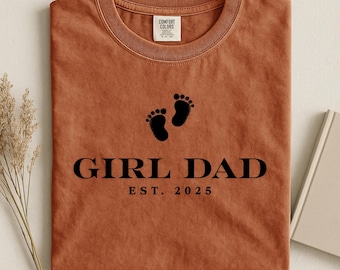 May include: A rust-colored t-shirt with a crew neck. The shirt features black text that reads "GIRL DAD EST. 2025" below two black footprints. The shirt has a tag that reads "COMFORT COLORS".