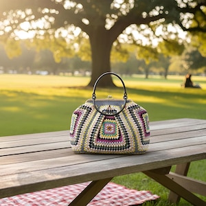 May include: A colorful, crocheted handbag with a black handle sits on a wooden picnic table. The bag features a geometric pattern with shades of purple, yellow, green, and gray. The background shows a park with a large tree and a person sitting on the grass.