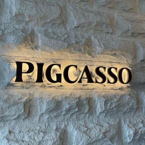Puede incluir: Un letrero de pared con el texto PIGCASSO en letras negras may&uacute;sculas. Las letras est&aacute;n retroiluminadas con una luz c&aacute;lida y brillante, creando una silueta definida sobre la superficie de una pared de bloques de piedra gris, rugosa y texturizada.