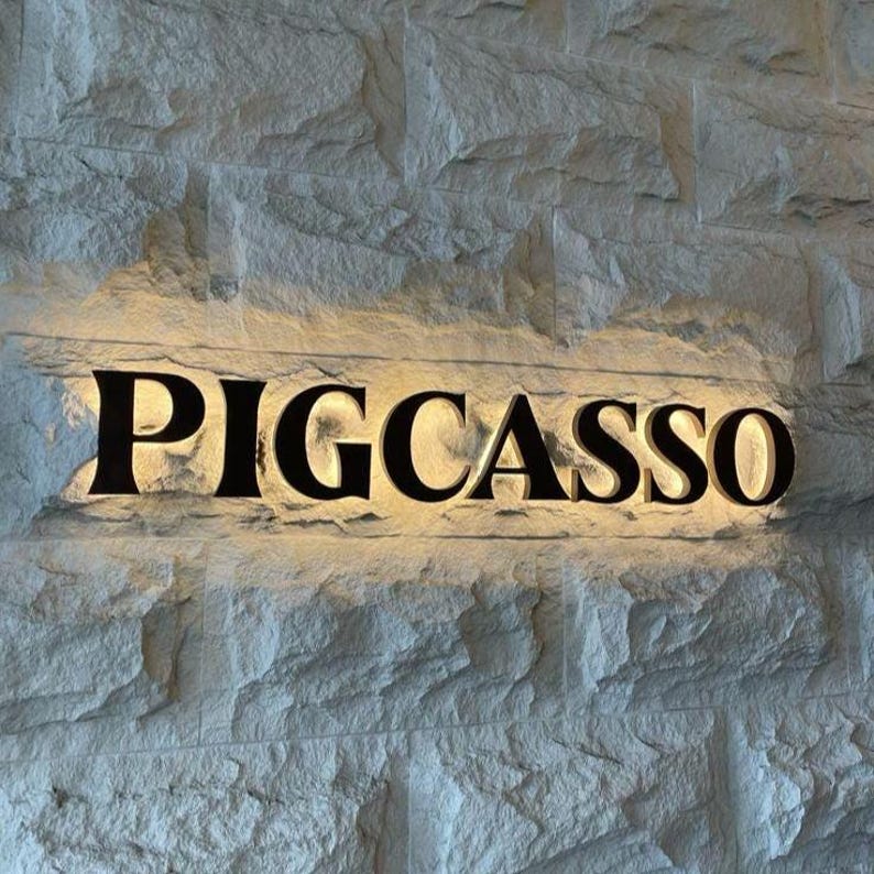 Puede incluir: Un letrero de pared con el texto PIGCASSO en letras negras may&uacute;sculas. Las letras est&aacute;n retroiluminadas con una luz c&aacute;lida y brillante, creando una silueta definida sobre la superficie de una pared de bloques de piedra gris, rugosa y texturizada.