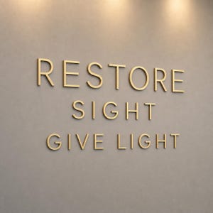 Puede incluir: Una decoración de pared con letras tridimensionales de color dorado dispuestas en tres filas. El texto dice RESTORE en la línea superior, SIGHT en la línea central y GIVE LIGHT en la línea inferior, sobre una pared de tonos neutros.