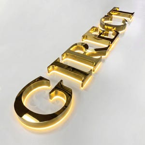 Puede incluir: Un conjunto de letras de metal dorado en tres dimensiones que forman la palabra GIRBOL, con una superficie reflectante pulida y retroiluminaci&oacute;n amarilla c&aacute;lida integrada que proyecta un suave resplandor alrededor de cada car&aacute;cter.