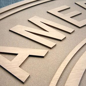 Puede incluir: Un letrero de madera de color marr&oacute;n claro con letras en relieve cortadas con l&aacute;ser que forman la palabra AMER. La superficie tiene un acabado mate suave con veta de madera visible, y las letras est&aacute;n elevadas, creando un efecto 3D.