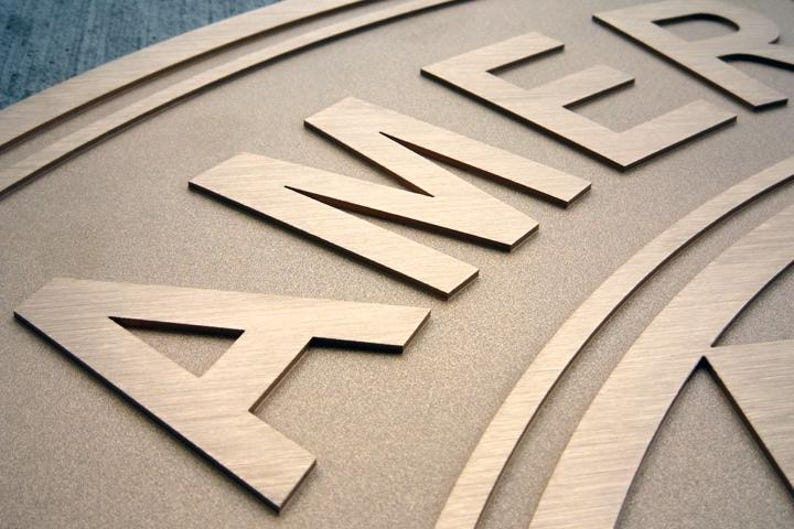 Puede incluir: Un letrero de madera de color marr&oacute;n claro con letras en relieve cortadas con l&aacute;ser que forman la palabra AMER. La superficie tiene un acabado mate suave con veta de madera visible, y las letras est&aacute;n elevadas, creando un efecto 3D.