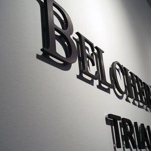 Puede incluir: Letras negras tridimensionales montadas en una pared, que deletrean BELCHER LANG y TRIAL ATTORNEYS. Las letras tienen un acabado met&aacute;lico y proyectan sombras marcadas sobre la superficie clara, creando un efecto de profundidad en la se&ntilde;alizaci&oacute;n.