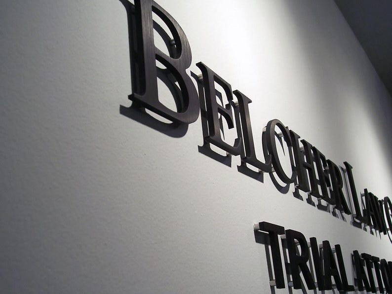 Puede incluir: Letras negras tridimensionales montadas en una pared, que deletrean BELCHER LANG y TRIAL ATTORNEYS. Las letras tienen un acabado met&aacute;lico y proyectan sombras marcadas sobre la superficie clara, creando un efecto de profundidad en la se&ntilde;alizaci&oacute;n.