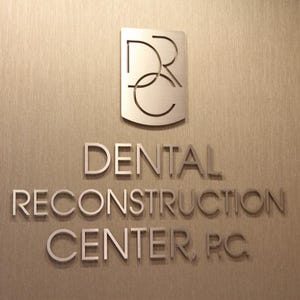 Puede incluir: Un letrero de pared en tono plateado que presenta las letras DRC dentro de un emblema en forma de escudo sobre el texto DENTAL RECONSTRUCTION CENTER, P.C. Las letras y el logotipo met&aacute;licos est&aacute;n montados en una pared texturizada de color marr&oacute;n claro.