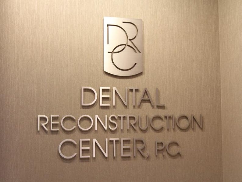 Puede incluir: Un letrero de pared en tono plateado que presenta las letras DRC dentro de un emblema en forma de escudo sobre el texto DENTAL RECONSTRUCTION CENTER, P.C. Las letras y el logotipo met&aacute;licos est&aacute;n montados en una pared texturizada de color marr&oacute;n claro.