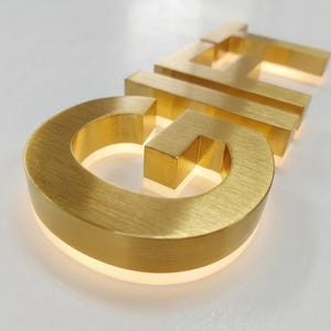 Puede incluir: Una letra G tridimensional fabricada en metal dorado cepillado con un acabado met&aacute;lico liso. La letra presenta un dise&ntilde;o hueco en la parte posterior que emite un suave resplandor blanco c&aacute;lido desde abajo, creando un efecto de halo.