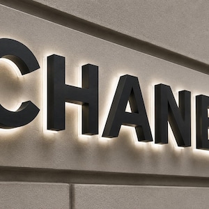 Puede incluir: Un letrero tridimensional que muestra la palabra CHANEL en letras may&uacute;sculas negras y negritas. Cada letra est&aacute; montada ligeramente separada de una pared de paneles de color beige claro, creando un halo blanco brillante tras los caracteres.