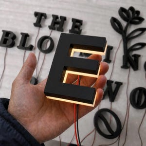 Puede incluir: Una letra E tridimensional con acabado negro mate y retroiluminaci&oacute;n LED blanca c&aacute;lida integrada. La letra presenta un centro hueco y dos cables el&eacute;ctricos delgados que salen de la parte inferior, con otras letras negras visibles detr&aacute;s.