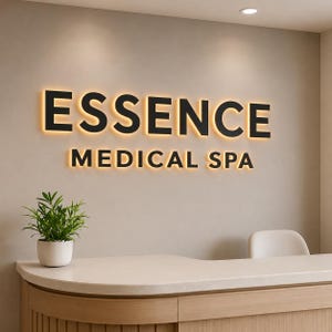 Puede incluir: Un letrero de pared con el texto negro ESSENCE MEDICAL SPA con retroiluminaci&oacute;n c&aacute;lida. El letrero est&aacute; colocado sobre un mostrador de recepci&oacute;n de color claro con una peque&ntilde;a planta verde en maceta a la izquierda y una silla blanca a la derecha.