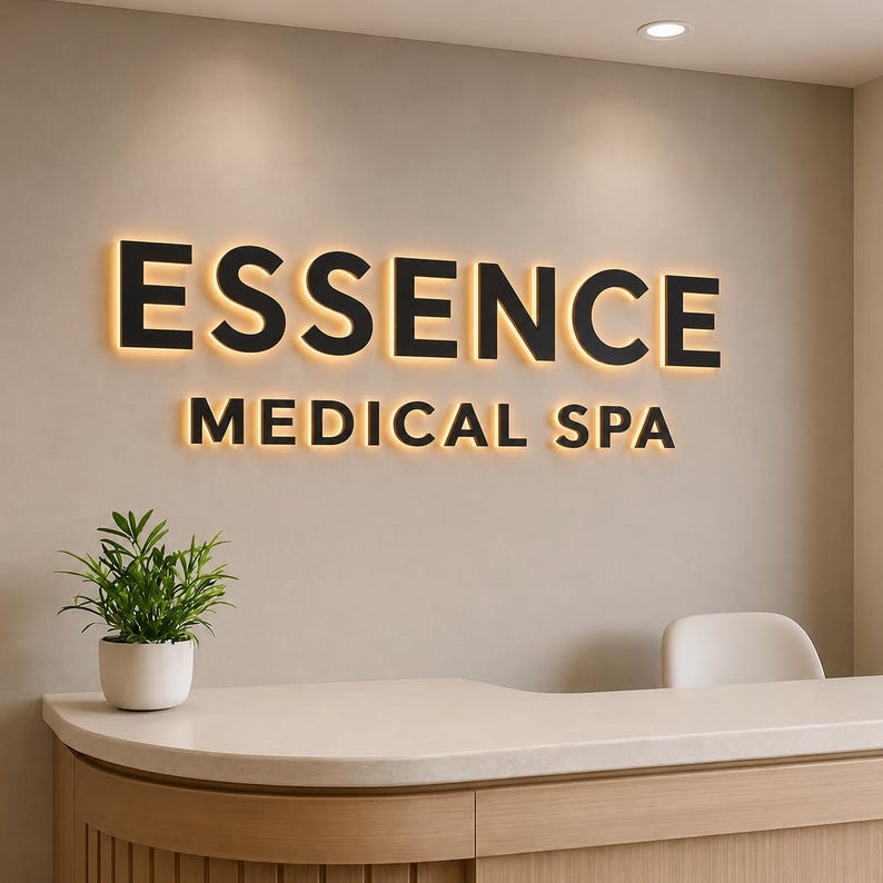 Puede incluir: Un letrero de pared con el texto negro ESSENCE MEDICAL SPA con retroiluminaci&oacute;n c&aacute;lida. El letrero est&aacute; colocado sobre un mostrador de recepci&oacute;n de color claro con una peque&ntilde;a planta verde en maceta a la izquierda y una silla blanca a la derecha.