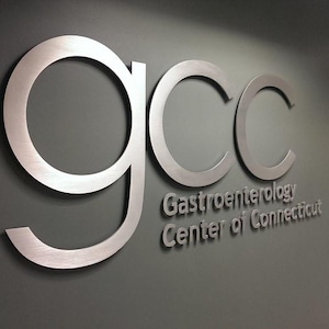 Puede incluir: Un letrero de pared tridimensional con las letras min&uacute;sculas gcc en un acabado plateado cepillado. Debajo de las letras, el texto Gastroenterology Center of Connecticut se muestra en una fuente plateada a juego sobre una pared gris oscuro.