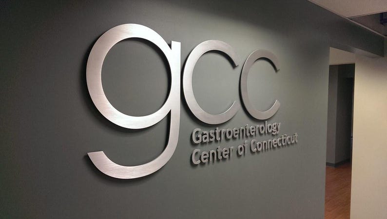 Puede incluir: Un letrero de pared tridimensional con las letras min&uacute;sculas gcc en un acabado plateado cepillado. Debajo de las letras, el texto Gastroenterology Center of Connecticut se muestra en una fuente plateada a juego sobre una pared gris oscuro.