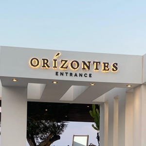 Puede incluir: Una estructura de entrada arquitect&oacute;nica blanca que presenta las letras tridimensionales retroiluminadas de color oscuro "ORIZONTES" con un peque&ntilde;o icono gr&aacute;fico sobre la letra I, y la palabra "ENTRANCE" en letras oscuras no iluminadas.