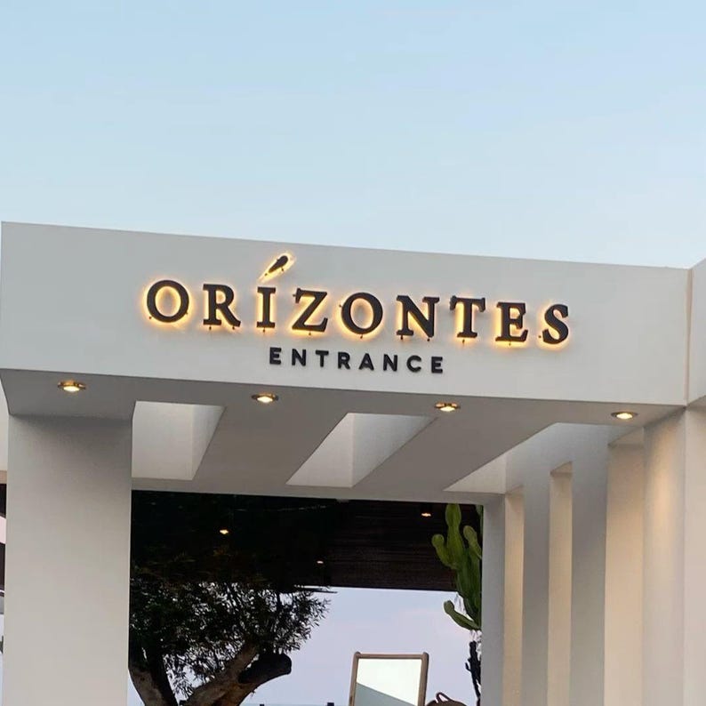 Puede incluir: Una estructura de entrada arquitect&oacute;nica blanca que presenta las letras tridimensionales retroiluminadas de color oscuro "ORIZONTES" con un peque&ntilde;o icono gr&aacute;fico sobre la letra I, y la palabra "ENTRANCE" en letras oscuras no iluminadas.