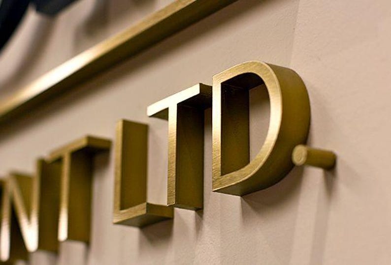 Puede incluir: Letras de metal de color dorado en tres dimensiones que forman la palabra LTD con un punto al final. Las letras tienen un acabado cepillado y est&aacute;n montadas sobre una superficie plana, creando un efecto de sombra distintivo y profesional.