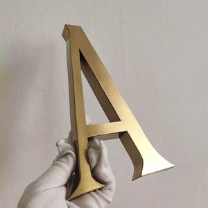 Puede incluir: Una letra may&uacute;scula A tridimensional hecha de metal cepillado de color dorado. La letra presenta una tipograf&iacute;a con remates, bordes limpios y afilados, y un acabado met&aacute;lico liso que refleja la luz en su parte frontal y en sus lados.