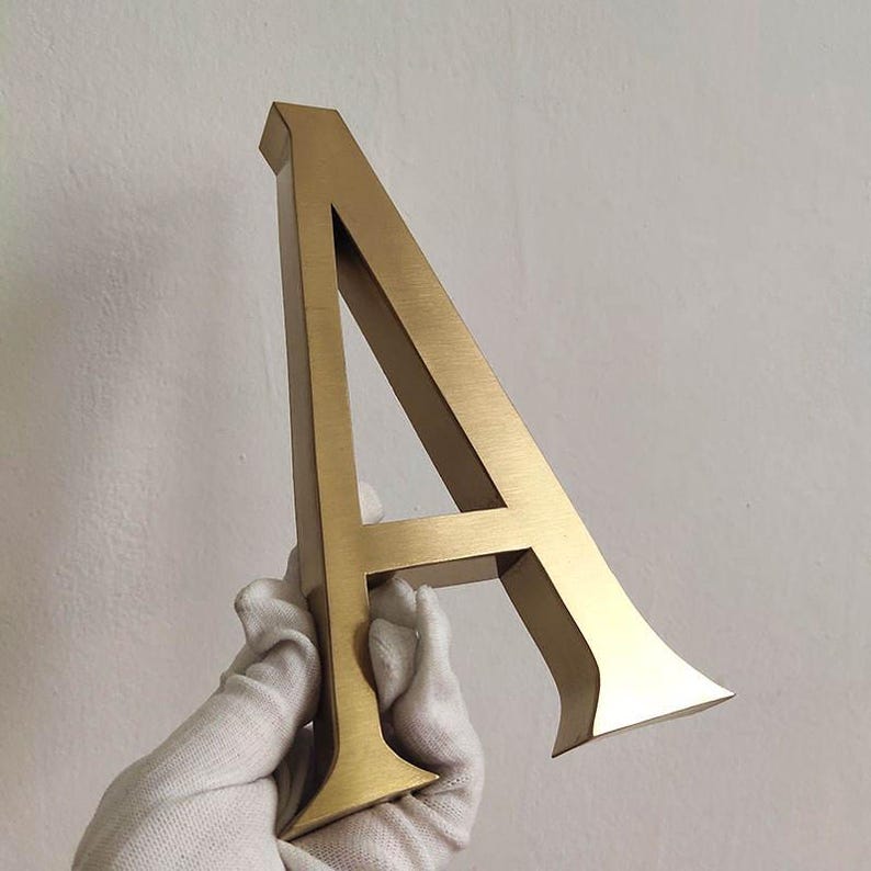 Puede incluir: Una letra may&uacute;scula A tridimensional hecha de metal cepillado de color dorado. La letra presenta una tipograf&iacute;a con remates, bordes limpios y afilados, y un acabado met&aacute;lico liso que refleja la luz en su parte frontal y en sus lados.