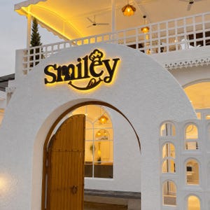 Puede incluir: Un edificio blanco con una entrada arqueada, una puerta de madera y un letrero negro retroiluminado que dice Smiley. La estructura incluye una barandilla de balc&oacute;n de celos&iacute;a blanca, iluminaci&oacute;n amarilla c&aacute;lida y un acabado de estuco texturizado.