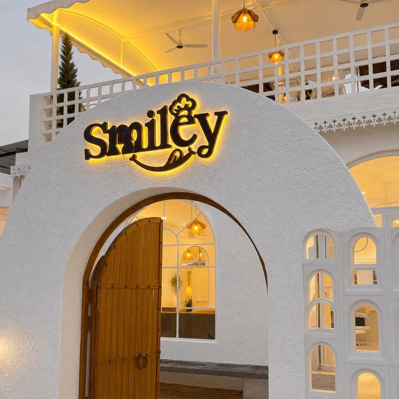Puede incluir: Un edificio blanco con una entrada arqueada, una puerta de madera y un letrero negro retroiluminado que dice Smiley. La estructura incluye una barandilla de balc&oacute;n de celos&iacute;a blanca, iluminaci&oacute;n amarilla c&aacute;lida y un acabado de estuco texturizado.