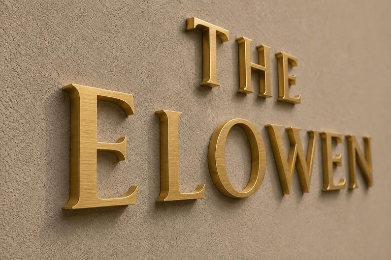 Puede incluir: Letras de metal de color dorado en tres dimensiones que forman la palabra THE ELOWEN. Las letras tienen un acabado cepillado y est&aacute;n montadas sobre una superficie texturizada de color beige claro, creando un efecto de sombra bajo cada car&aacute;cter.