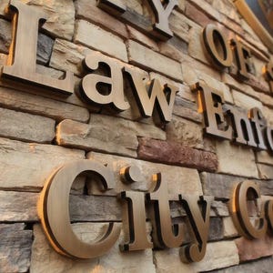 Puede incluir: Un muro de piedra con letras met&aacute;licas tridimensionales de color bronce que forman la inscripci&oacute;n City of Law Enforcement City Council. El muro est&aacute; construido con piedras rectangulares apiladas en tonos beige, gris y marr&oacute;n.