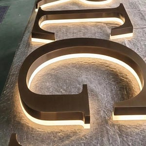 Puede incluir: Un conjunto de letras met&aacute;licas tridimensionales con acabado en bronce cepillado, que cuentan con retroiluminaci&oacute;n LED blanca c&aacute;lida integrada que ilumina los bordes. Las letras est&aacute;n dispuestas en secuencia, con la letra C en primer plano.
