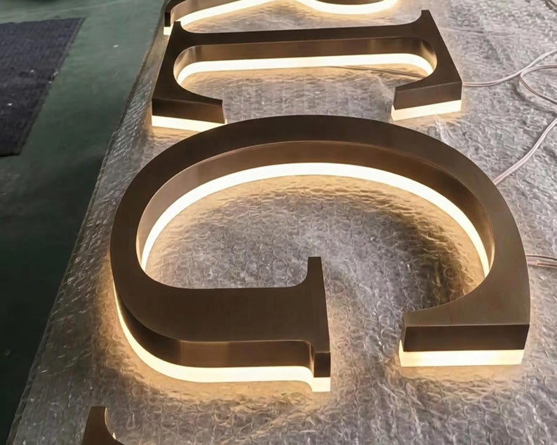 Puede incluir: Un conjunto de letras met&aacute;licas tridimensionales con acabado en bronce cepillado, que cuentan con retroiluminaci&oacute;n LED blanca c&aacute;lida integrada que ilumina los bordes. Las letras est&aacute;n dispuestas en secuencia, con la letra C en primer plano.