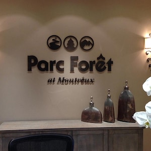 Puede incluir: Una exhibici&oacute;n de pared con el texto de metal oscuro Parc For&ecirc;t at Montr&ecirc;ux y tres iconos circulares encima. Dos apliques de pared con pantallas blancas flanquean el letrero. Tres vasijas decorativas met&aacute;licas sobre un mueble de madera.