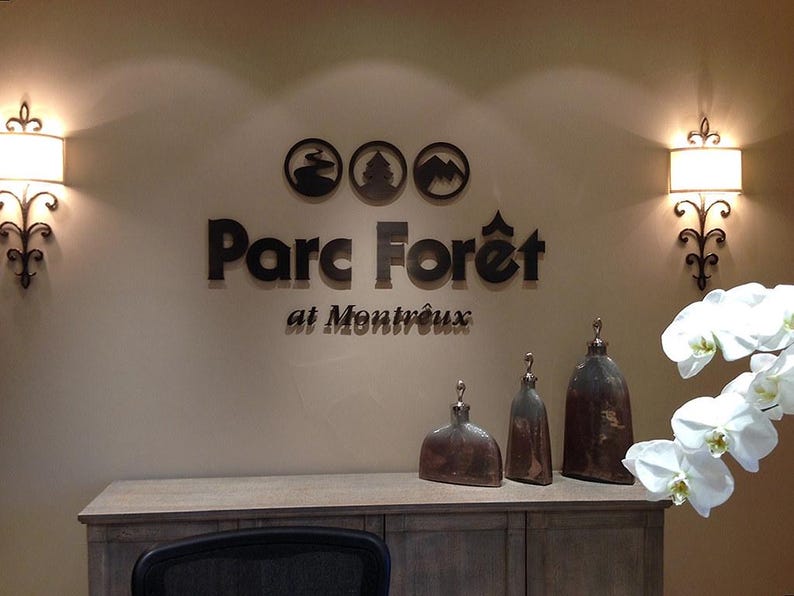 Puede incluir: Una exhibici&oacute;n de pared con el texto de metal oscuro Parc For&ecirc;t at Montr&ecirc;ux y tres iconos circulares encima. Dos apliques de pared con pantallas blancas flanquean el letrero. Tres vasijas decorativas met&aacute;licas sobre un mueble de madera.