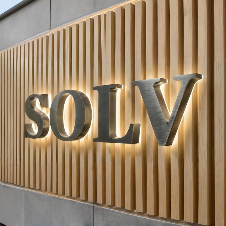 Puede incluir: Un letrero de pared con las letras SOLV en acabado met&aacute;lico cepillado, montado sobre un fondo de listones de madera clara verticales. Las letras est&aacute;n retroiluminadas, creando un efecto brillante sobre el panel de madera.