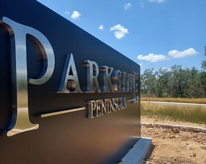 Puede incluir: Un letrero exterior grande, rectangular y de color negro mate con letras met&aacute;licas plateadas en relieve que forman la palabra PARKSIDE PENINSULA. El letrero est&aacute; montado sobre una base de hormig&oacute;n en un entorno exterior con hierba.