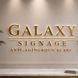 Puede incluir: Un letrero de pared con letras doradas en 3D que dice GALAXY SIGNAGE ANTI-AGING&QUICKCARE. El letrero est&aacute; iluminado con un brillo c&aacute;lido sobre una pared de tonos neutros, colocado sobre un mostrador de m&aacute;rmol blanco con una planta a cada lado.