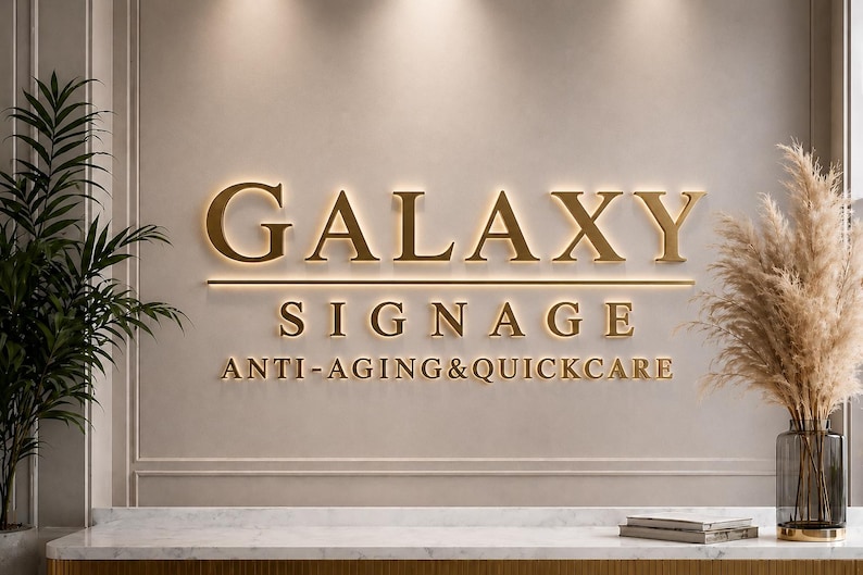 Puede incluir: Un letrero de pared con letras doradas en 3D que dice GALAXY SIGNAGE ANTI-AGING&QUICKCARE. El letrero est&aacute; iluminado con un brillo c&aacute;lido sobre una pared de tonos neutros, colocado sobre un mostrador de m&aacute;rmol blanco con una planta a cada lado.