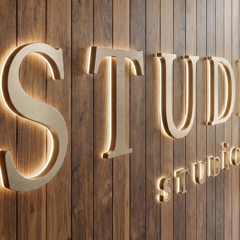 Puede incluir: Grandes letras tridimensionales de metal dorado cepillado que forman la palabra STUDIO, montadas sobre una pared vertical de listones de madera de color marr&oacute;n oscuro. Cada letra tiene una retroiluminaci&oacute;n LED c&aacute;lida.