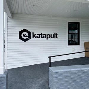 Puede incluir: El exterior de un edificio blanco con un logotipo negro en forma de hex&aacute;gono y el texto katapult. El edificio tiene revestimiento horizontal blanco, una ventana con marco negro, una base de ladrillo gris y una barandilla de metal negro.