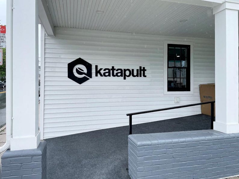 Puede incluir: El exterior de un edificio blanco con un logotipo negro en forma de hex&aacute;gono y el texto katapult. El edificio tiene revestimiento horizontal blanco, una ventana con marco negro, una base de ladrillo gris y una barandilla de metal negro.