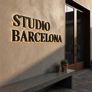 Puede incluir: Un letrero de pared con letras negras tridimensionales que dicen STUDIO BARCELONA, iluminadas desde atr&aacute;s con un brillo c&aacute;lido. El letrero est&aacute; montado en una pared beige texturizada junto a una puerta de cristal con marco oscuro y un banco de hormig&oacute;n.