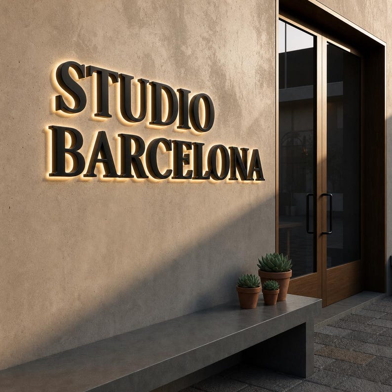Puede incluir: Un letrero de pared con letras negras tridimensionales que dicen STUDIO BARCELONA, iluminadas desde atr&aacute;s con un brillo c&aacute;lido. El letrero est&aacute; montado en una pared beige texturizada junto a una puerta de cristal con marco oscuro y un banco de hormig&oacute;n.